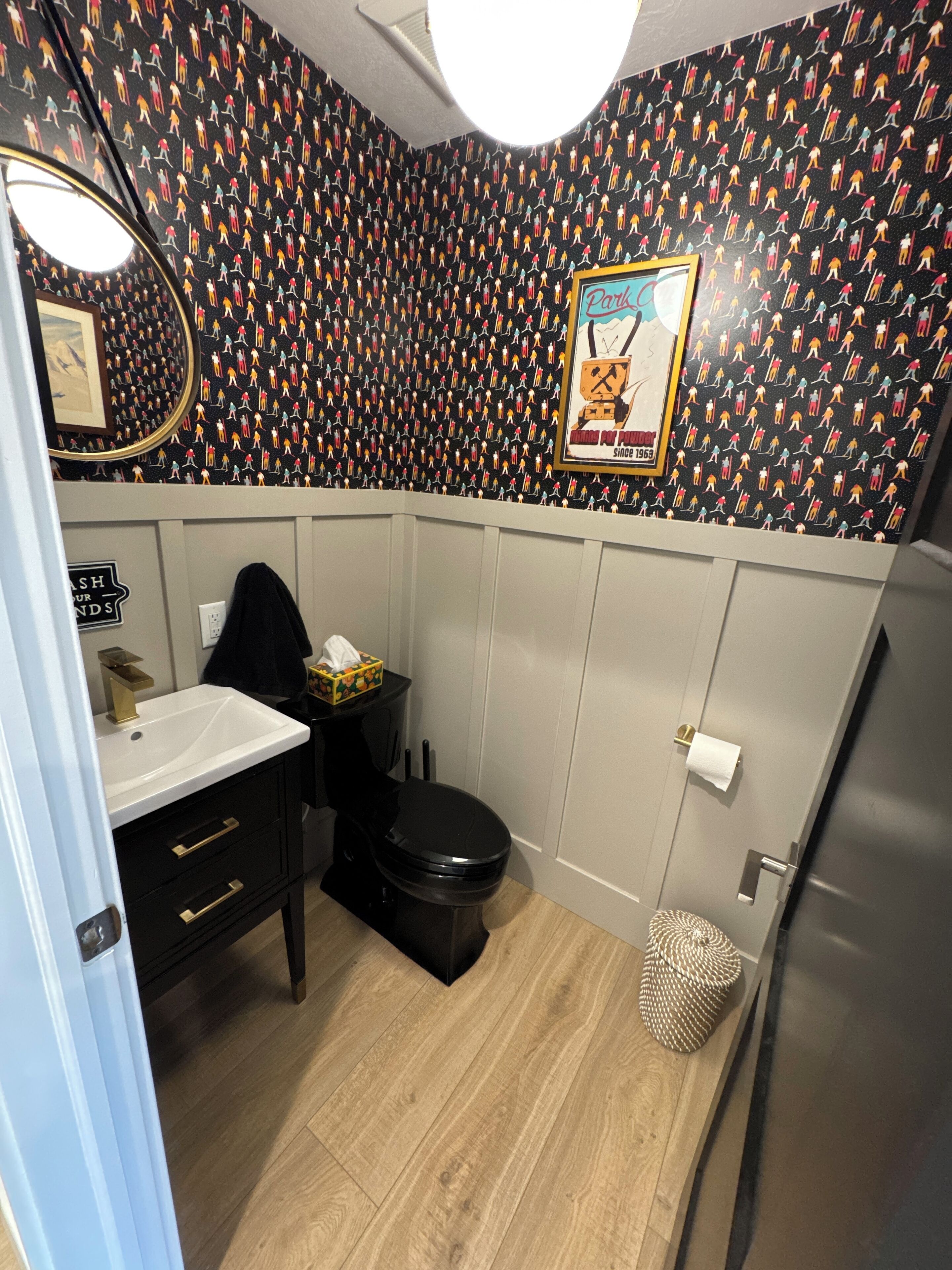Une salle de bain avec des toilettes noires, un lavabo blanc et un miroir. Il y a un tableau encadré au mur et une poubelle.