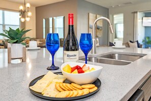 A modern kitchen with a countertop, a bottle of wine, two blue glasses, a bowl of fruit, cheese, and crackers.
