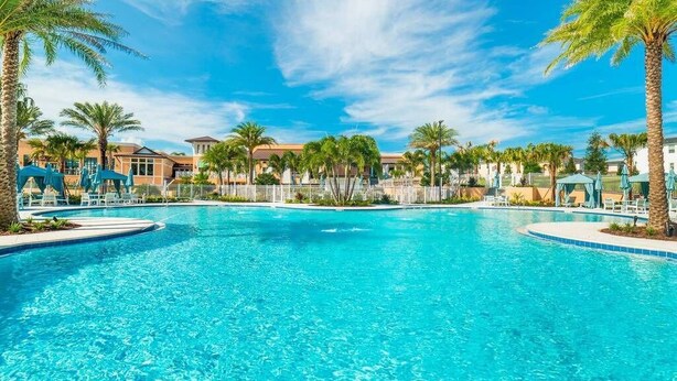 A large outdoor swimming pool surrounded by palm trees and lounge chairs, with buildings in the background. - Modern Decor With Themed Rooms, With Retro Game Room and Huge Pool in Upscale Resort! #6s820 (Kissimmee)