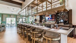 A modern bar area with a long counter, multiple stools, and a television screen displaying a news program. - Modern Decor With Themed Rooms, With Retro Game Room and Huge Pool in Upscale Resort! #6s820 (Kissimmee)
