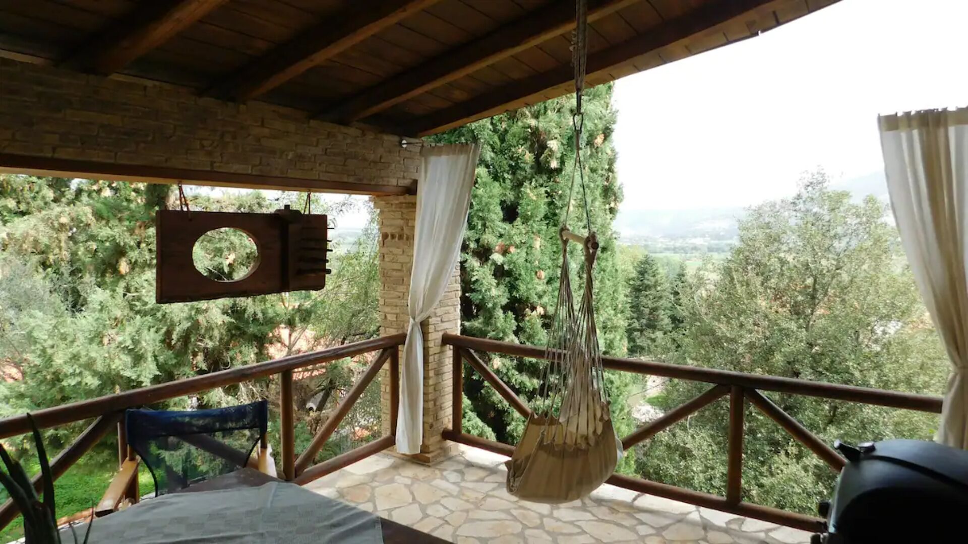 A balcony with a stone floor, a hanging chair, and a view of a forested landscape.