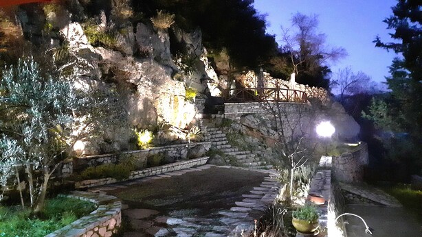 Un sendero de piedra que asciende por una ladera rocosa, rodeado de vegetación y un muro de piedra con barandilla de metal.