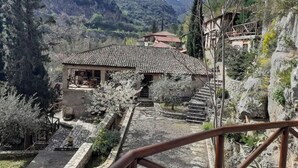 Un edificio de piedra tradicional con tejado de tejas, rodeado por un camino de piedra y árboles en flor, con montañas al fondo.