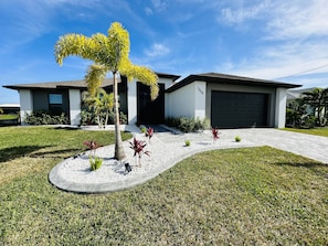 A modern two-story house with a garage, surrounded by a well-maintained lawn and tropical plants.