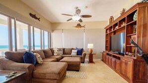 A living room with a large TV, wooden entertainment center, and a ceiling fan. - Ocean’s Edge 4B – Fourth-Floor Beachfront Condo with Wrap-Around Ocean Views (Ambergris Caye)