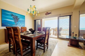 A dining area with a wooden table and chairs, a large painting of a fish, and a view of the ocean through a sliding glass door.