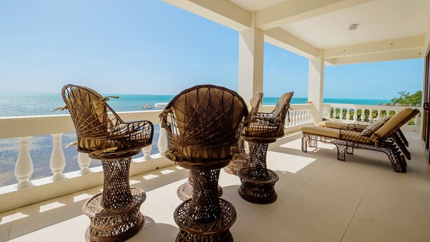 A covered outdoor seating area with wicker chairs and a lounge chair, overlooking a clear blue sea. - Ocean’s Edge 4B – Fourth-Floor Beachfront Condo with Wrap-Around Ocean Views (Ambergris Caye)