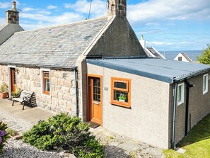 Une maison en pierre avec un toit gris, une porte en bois et une fenêtre ornée d’une plante.
