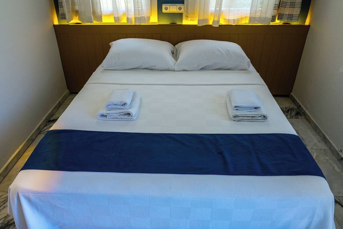 Una cama bien tendida con sábanas blancas y dos toallas dobladas, un cabecero de madera y una mesita de noche con una lámpara.