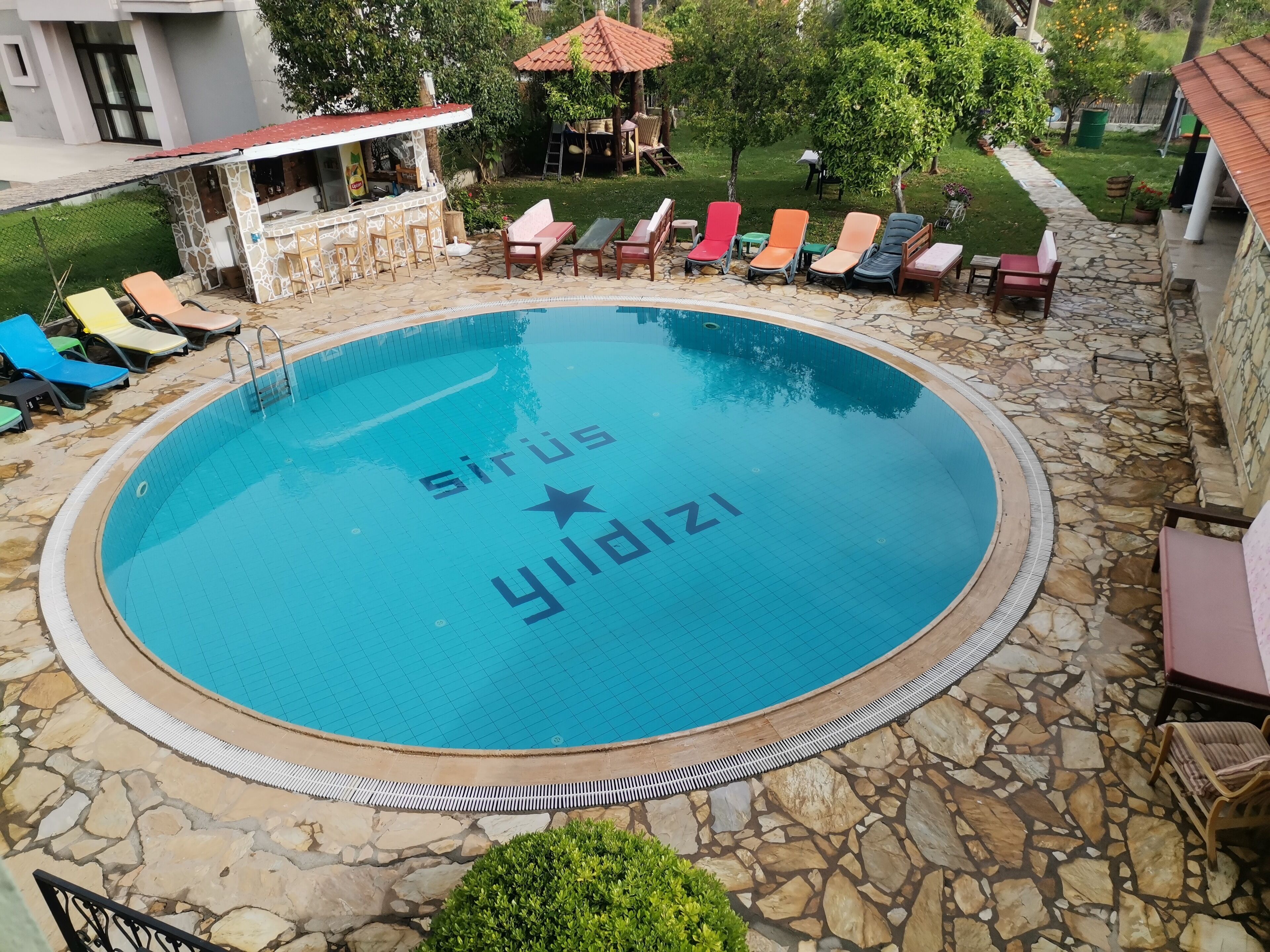 Una piscina circular con la inscripción 'Sirius Yildizi' en el agua, rodeada de baldosas de piedra y sillones de descanso.