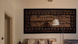 Un salon moderne avec une grande tapisserie décorative, un canapé agrémenté de coussins à motifs, une table basse avec un livre et des verres, et un lampadaire.