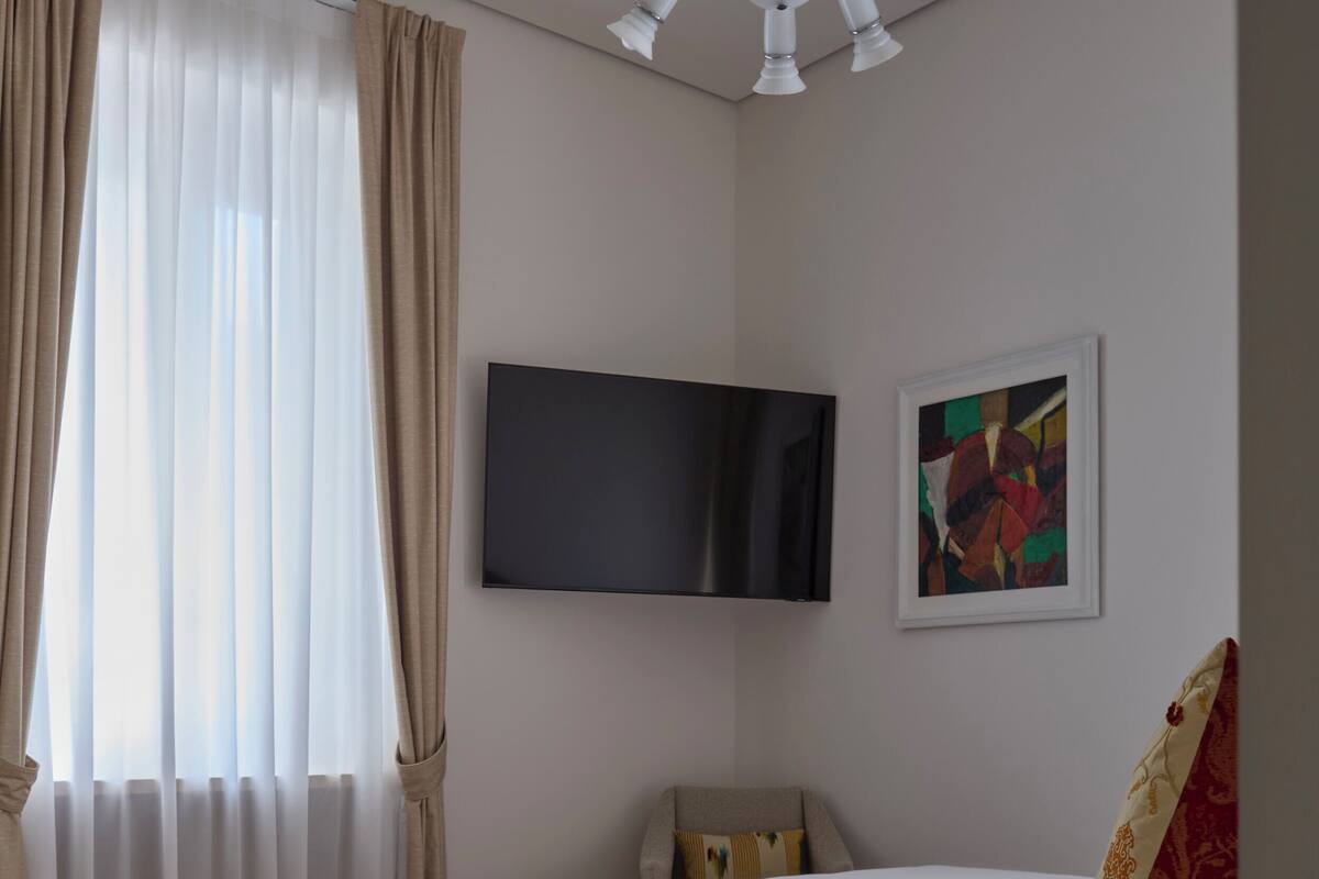 Habitación de hotel con una cama grande, un televisor de pantalla plana montado en la pared, un cuadro abstracto enmarco y una mesita de noche con un libro.