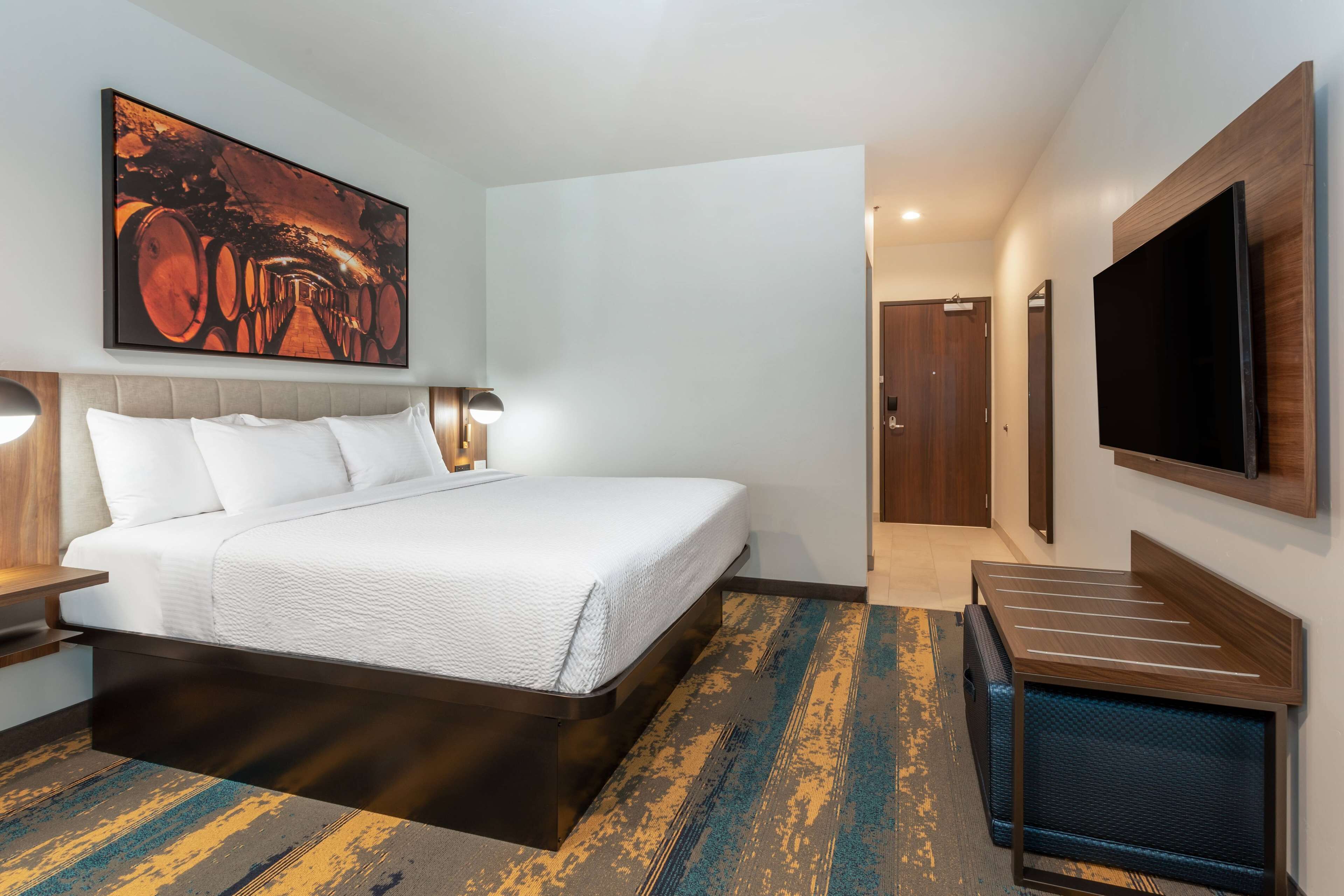 A modern hotel room with a large bed, a flat-screen TV, and a painting above the bed.