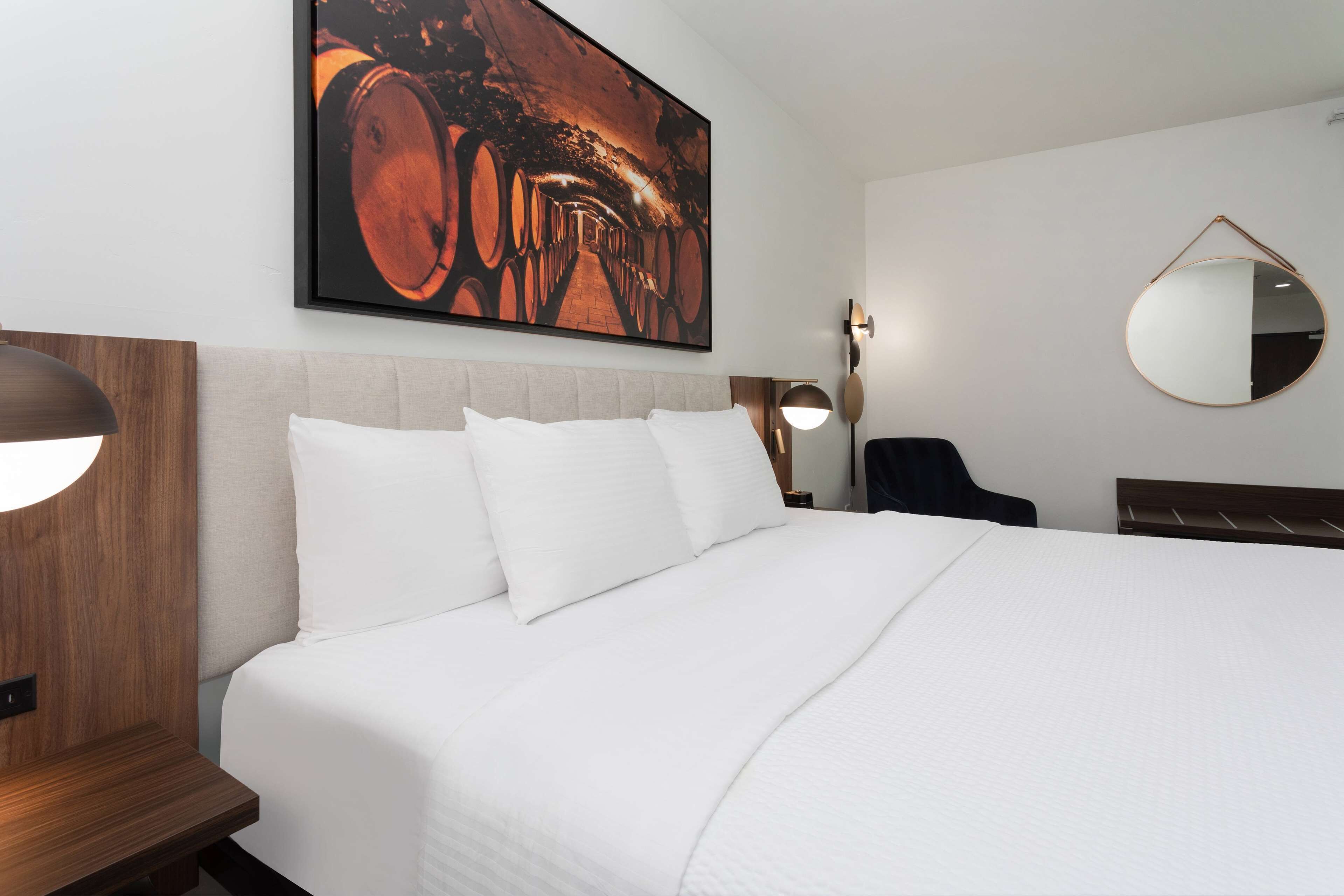 Habitación de hotel con una cama grande, cabecera, un espejo redondo y una obra de arte en la pared con barricas.