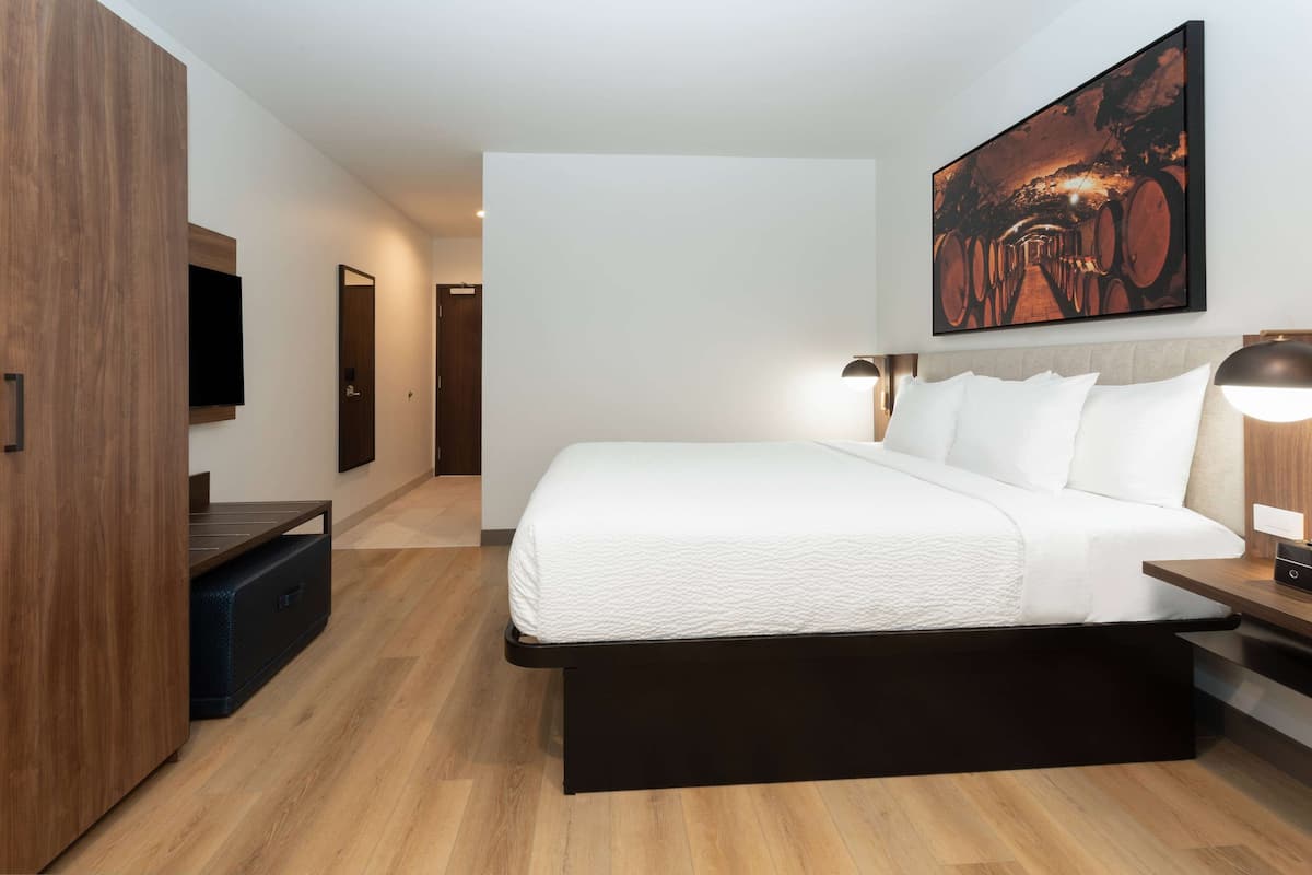 A modern hotel room with a large bed, a flat-screen TV, a wooden wardrobe, and a painting on the wall.