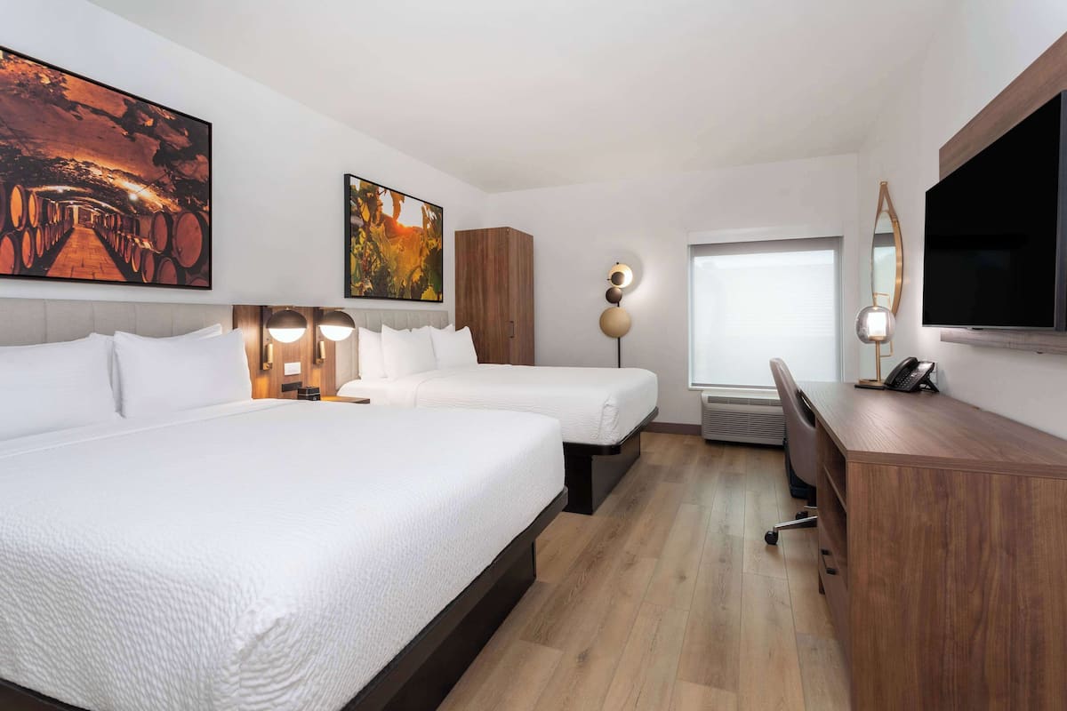 A hotel room with two beds, a desk, a chair, a TV, and artwork on the walls.