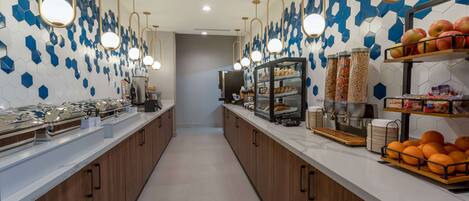 A long buffet line with food stations, including fruits and pastries, and a modern design with hexagonal tiles and pendant lighting.