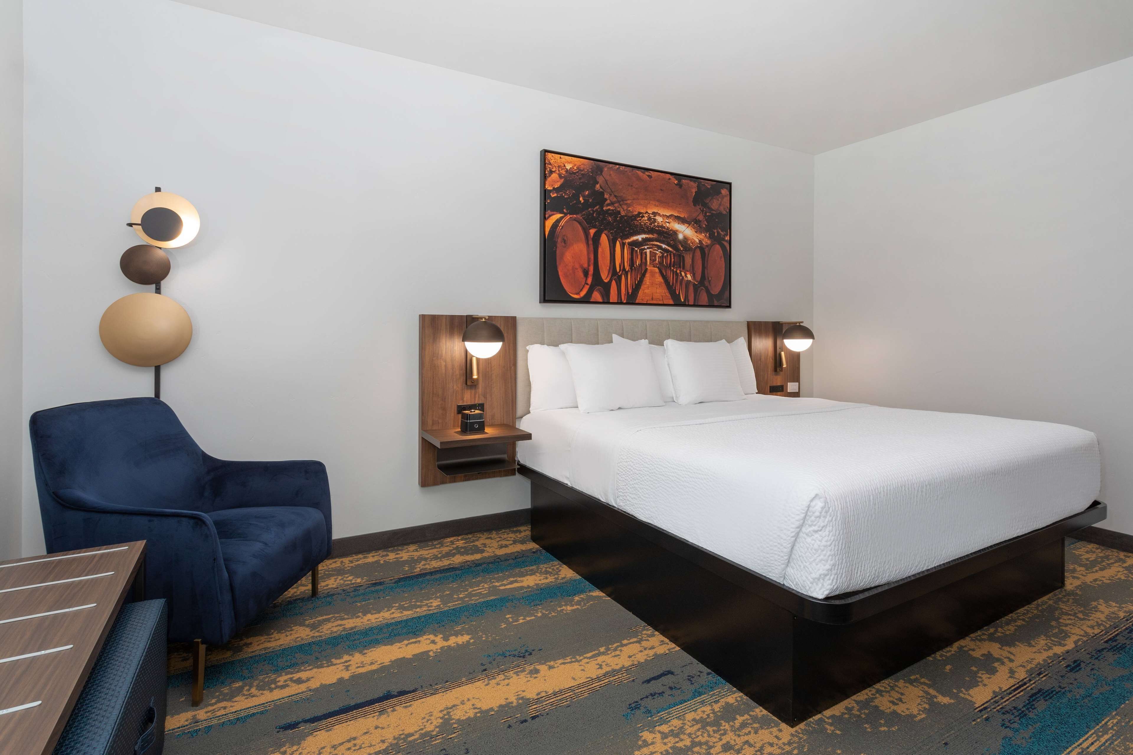 Una habitación de hotel con una cama grande, un sillón azul, un escritorio de madera y una obra de arte colgada en la pared.