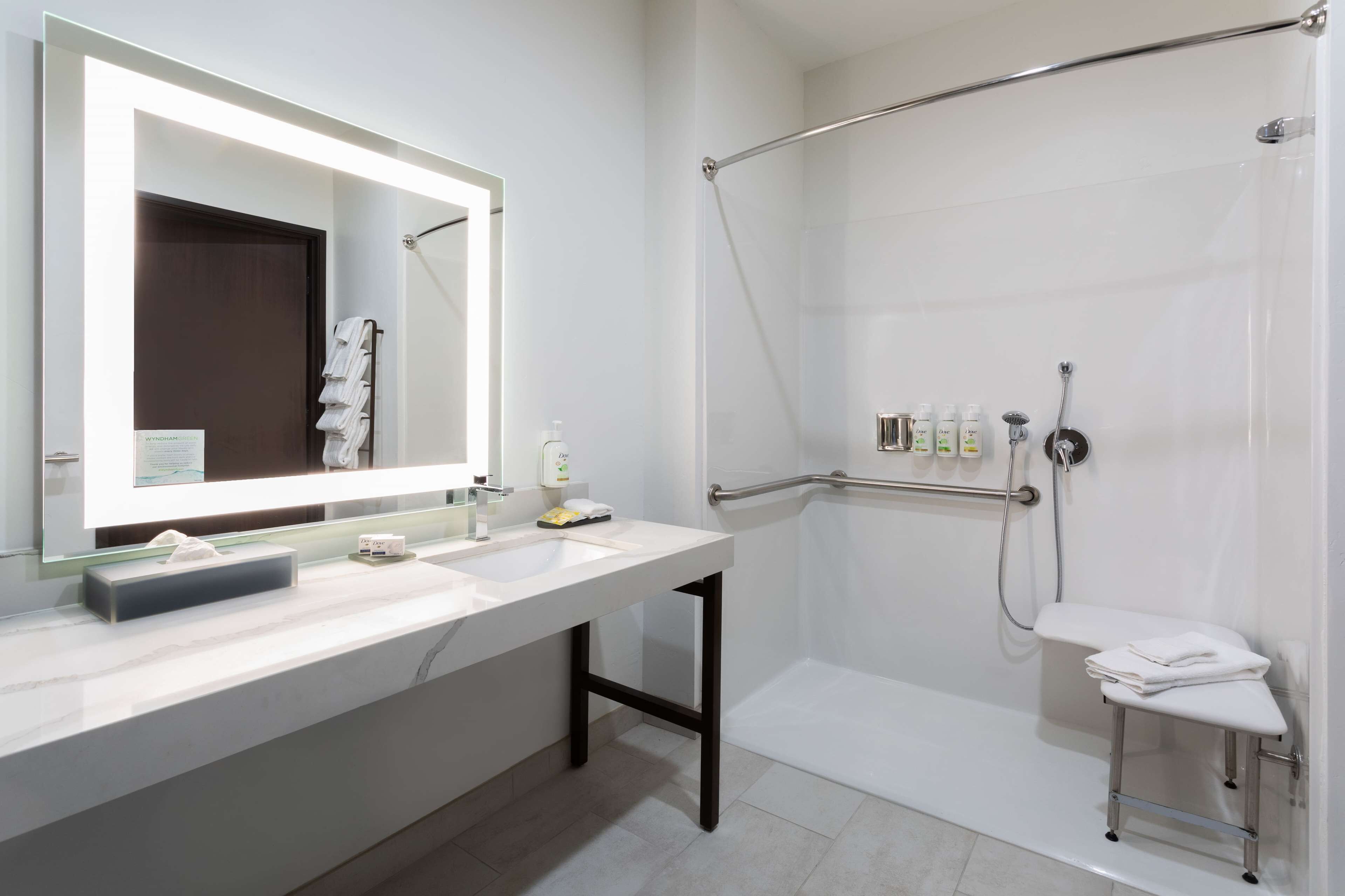 A modern bathroom with two sinks, a large mirror, a shower with a grab bar, and a small stool with towels.