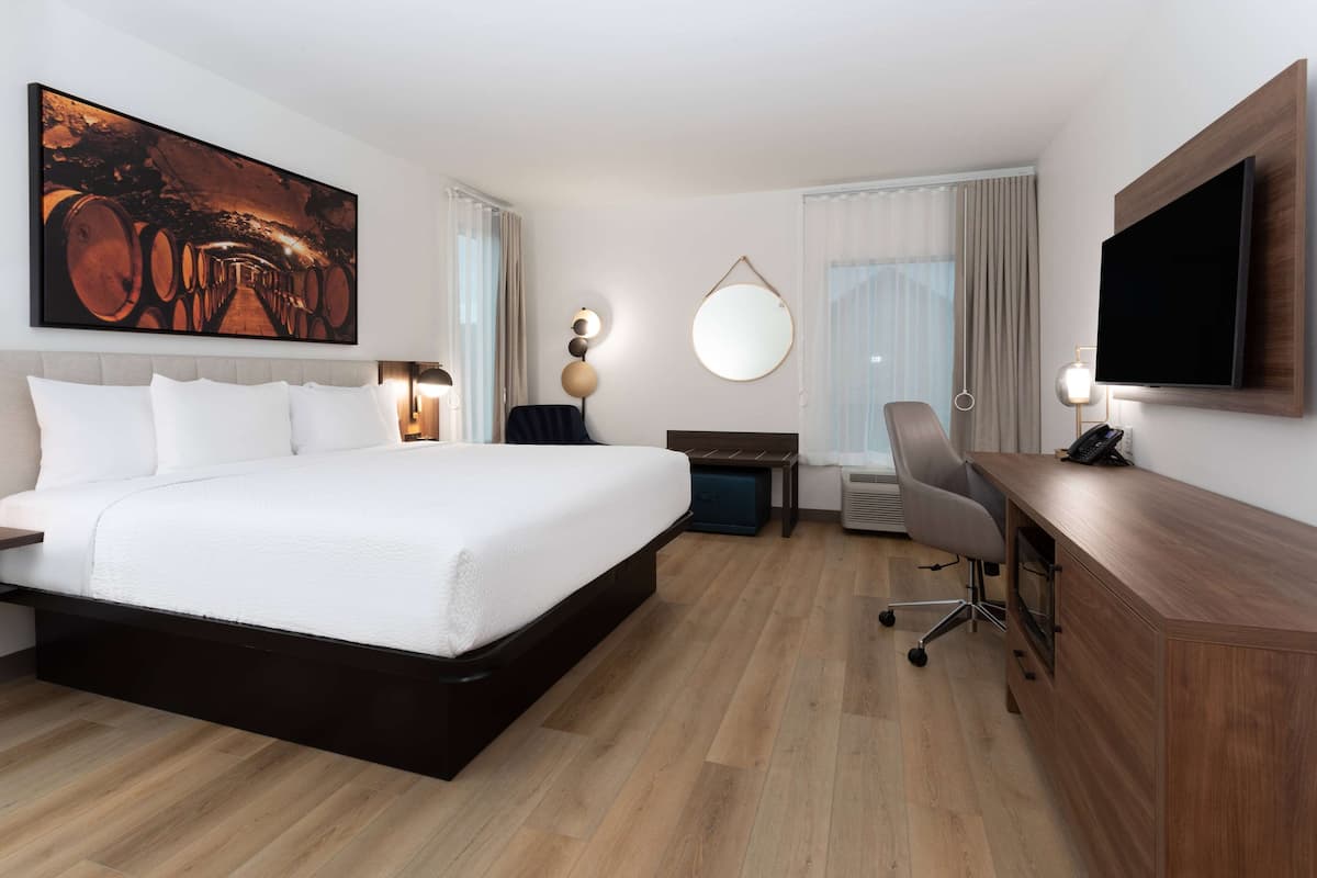 A hotel room with a large bed, a desk with a flat-screen TV, a round mirror, and a large artwork on the wall.