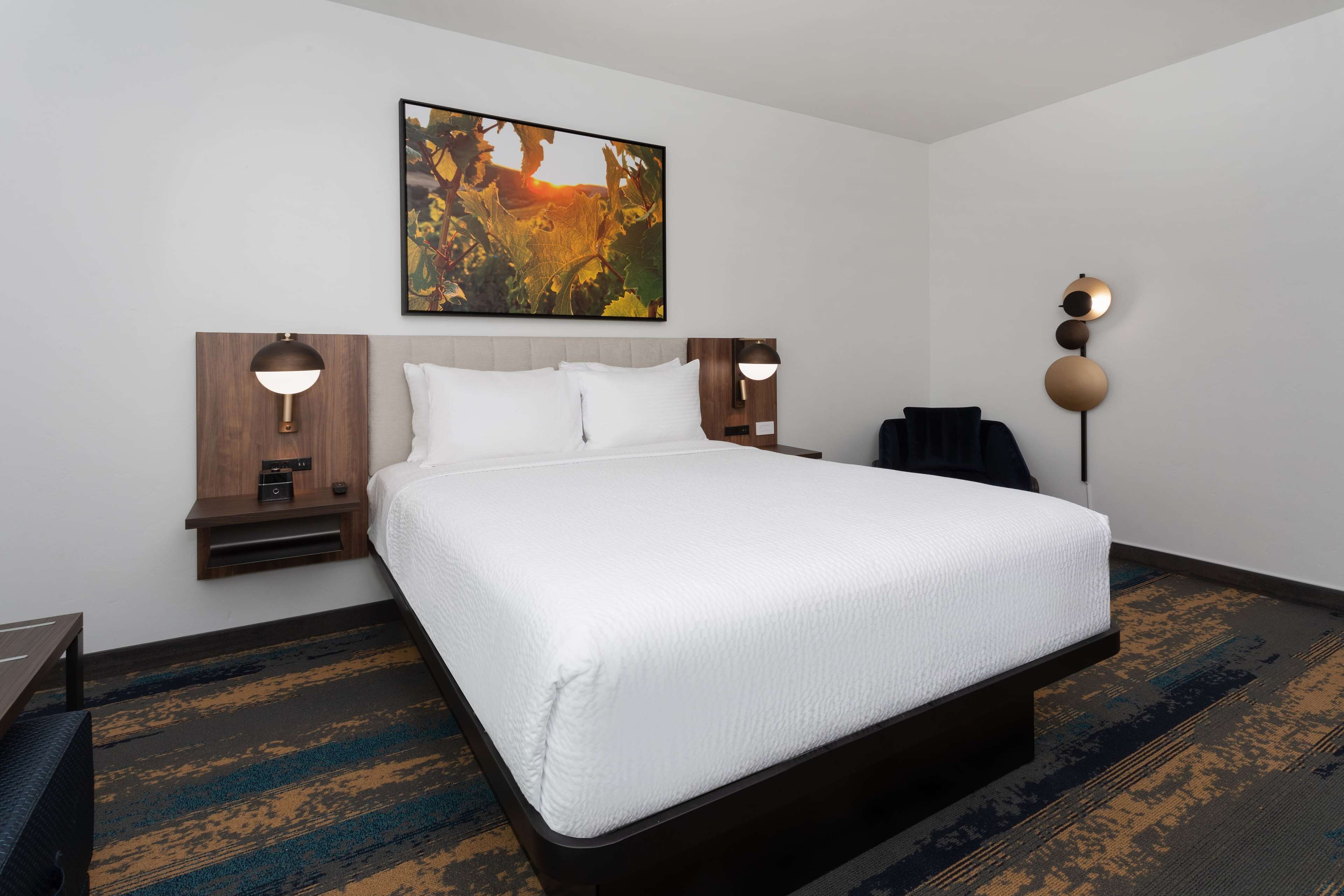 A hotel room with a large bed, two bedside tables with lamps, a wall-mounted artwork, and a patterned carpet.