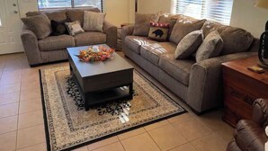 Un salon avec deux canapés, une table basse et un tapis. Il y a des fenêtres avec des stores, une porte et une lampe.