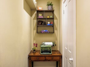 A narrow hallway with a wooden desk, a typewriter, and shelves holding various items including a potted plant and boxes.