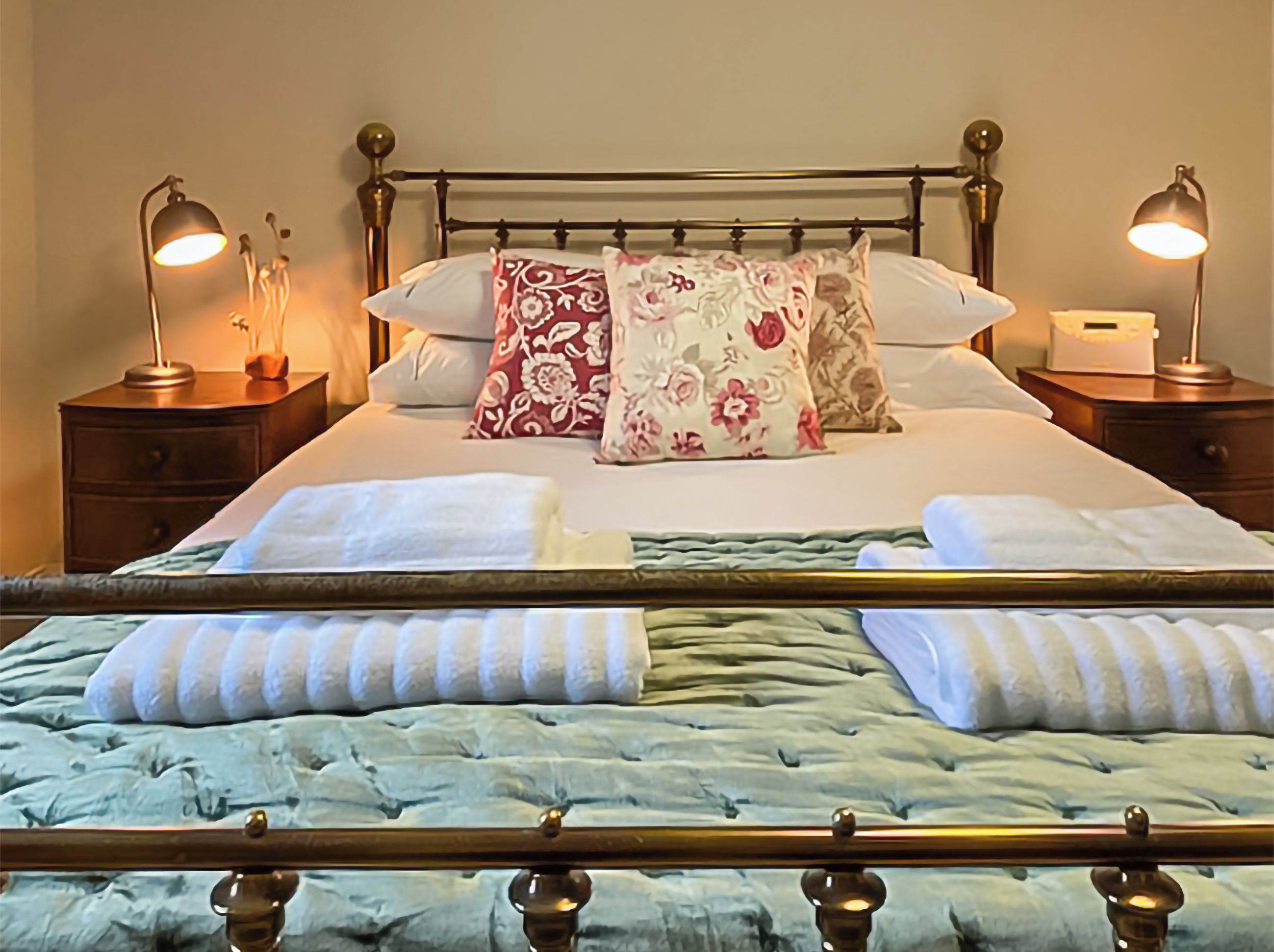Un lit bien fait, agrémenté d’oreillers à motifs floraux, une table de chevet avec une lampe et une petite table de nuit également dotée d’une lampe.