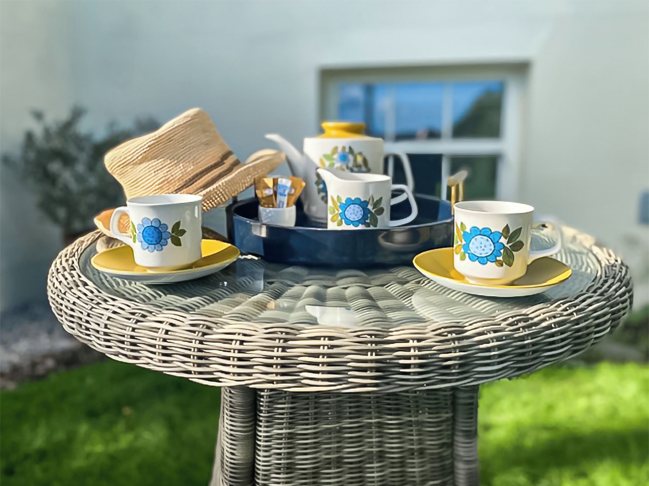 Ein Korbtisch mit zwei weißen Tassen mit blumigem, blauem Dekor, einem Strohhut und einer Tablett mit einer Kanne und Zuckerportionen.