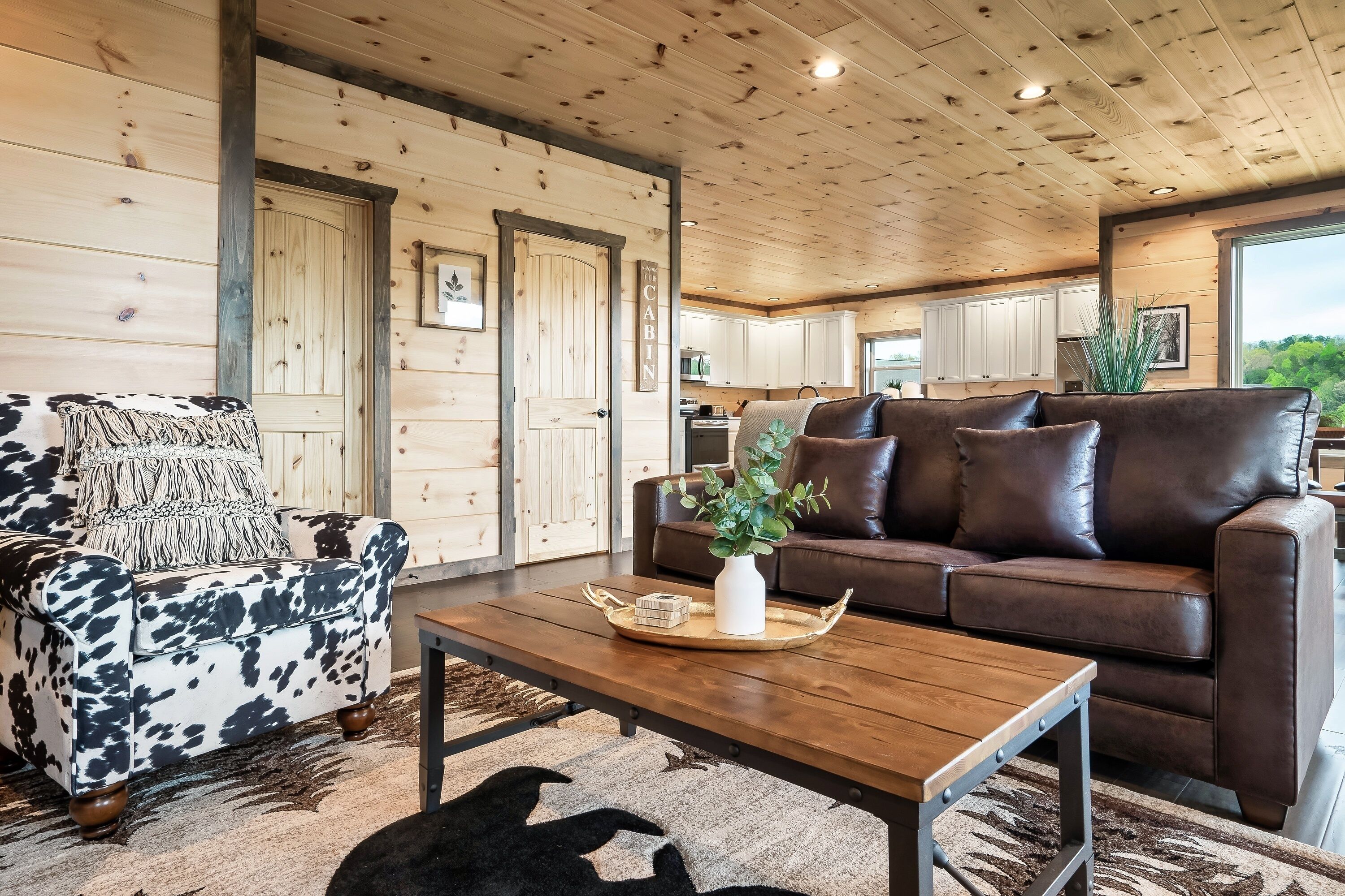 Un interior de cabaña con un sillón estampado con vacas, un sofá de cuero marrón, una mesa de centro de madera y una cocina al fondo.