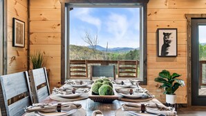 Un coin repas aménagé dans une cabane en bois, avec une table dressée pour six personnes, des œuvres d’art encadrées aux murs et une vue sur les montagnes.