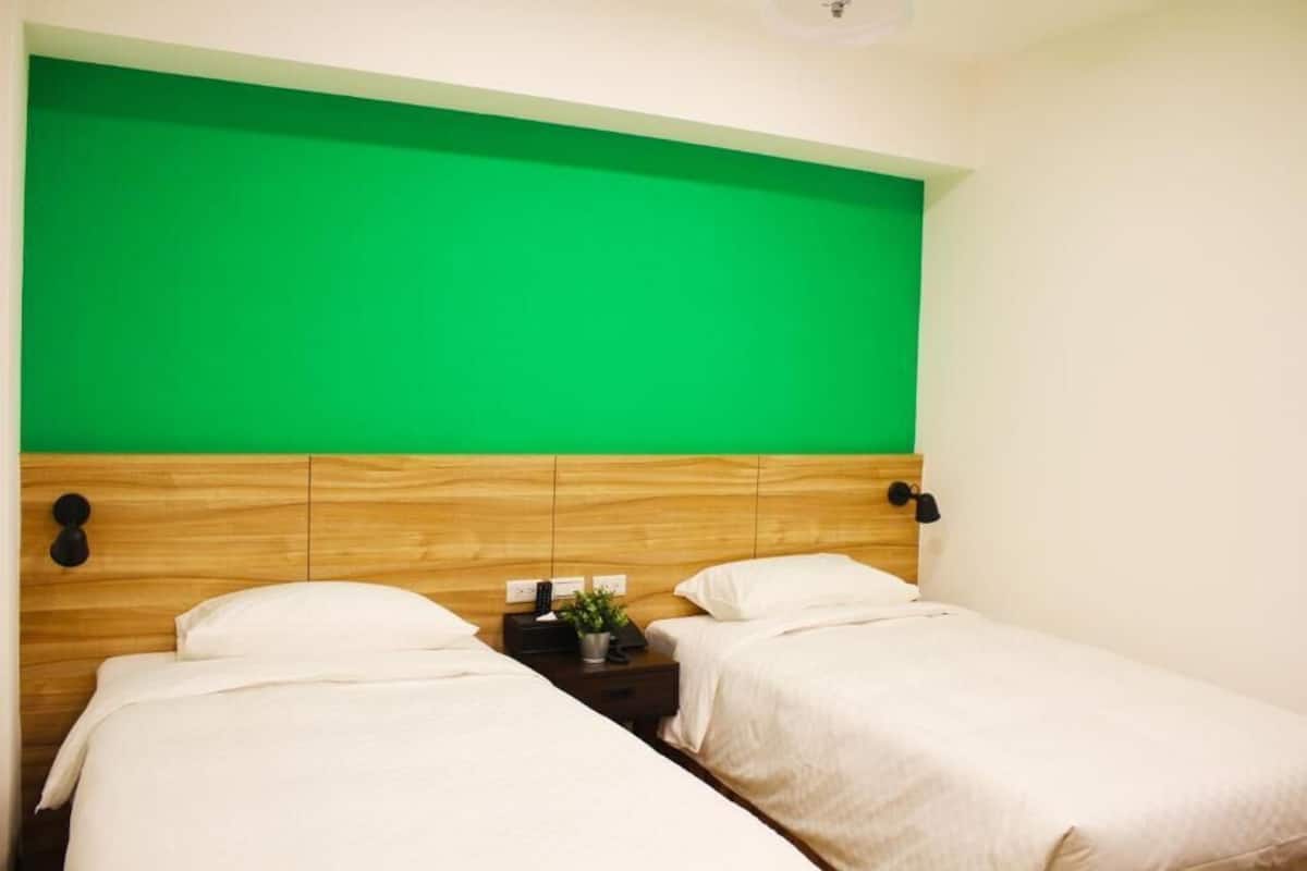 A room with two single beds, a wooden headboard, a small bedside table with a lamp, and a green screen above the beds.