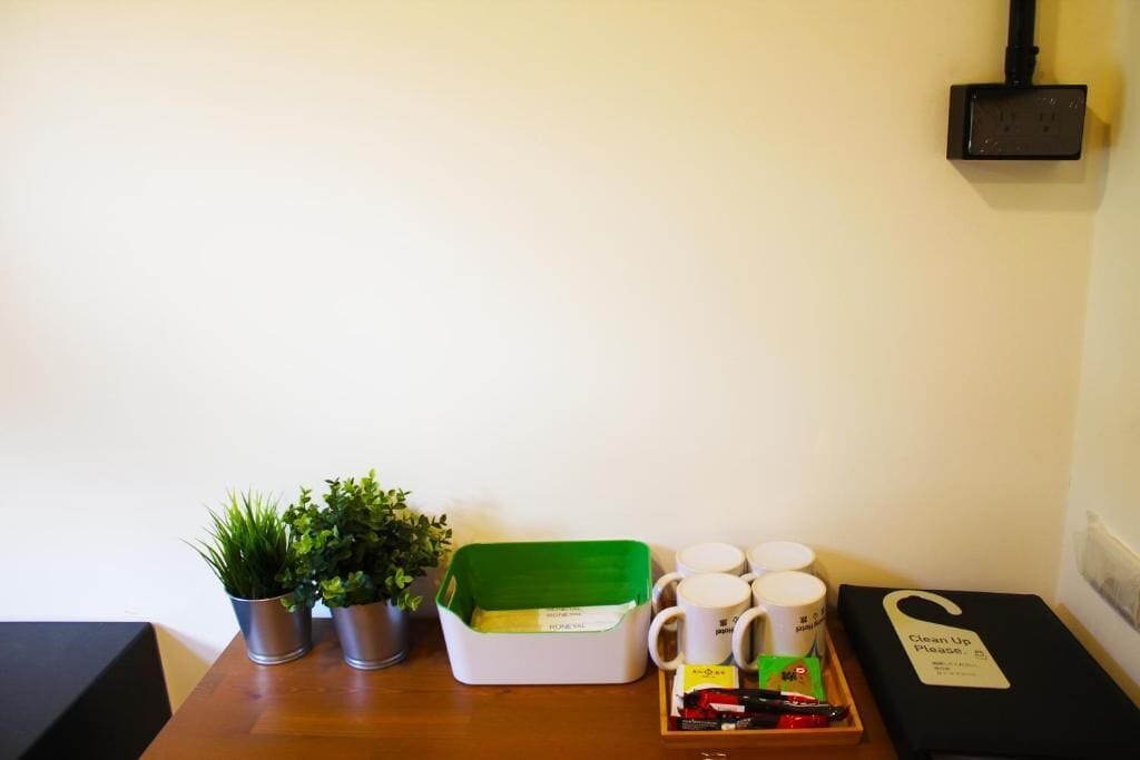 Une petite table avec un panier vert, des plantes en pot, un plateau avec des tasses et une pancarte indiquant « Veuillez ranger », ainsi qu’une boîte noire dotée d’une poignée.