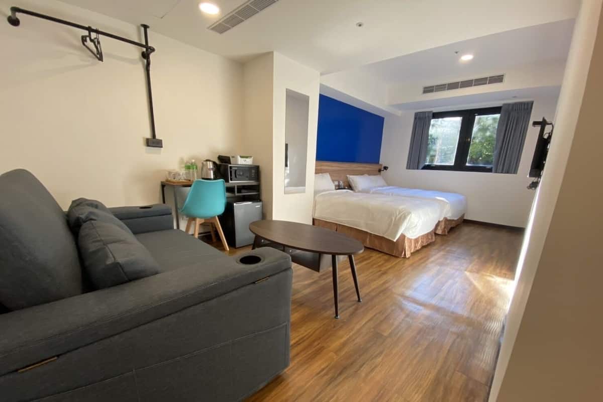 A modern hotel room with a sofa, a small table, a kitchenette, and a bed with two pillows.
