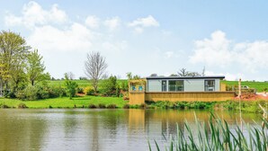 A lakeside cabin with a wooden deck, a small dock, and a grassy area with trees. - Ponsford Ponds - Goosedown Lodge (Knowle, near Cullompton)
