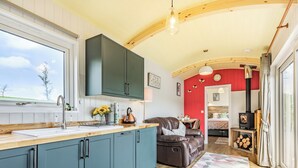 A kitchen with a large window, a sink, and a countertop. There is a dining area with a table and chairs, a sofa, and a red wall. A fireplace is visible in the background. - Ponsford Ponds - Goosedown Lodge (Knowle, near Cullompton)