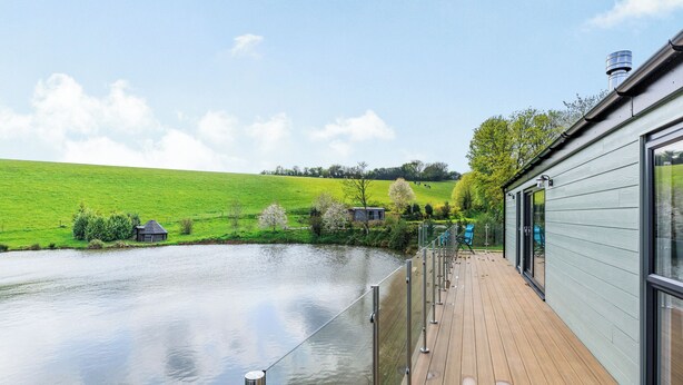 A wooden deck with a view of a river, a grassy field, and a building with large windows. - Ponsford Ponds - Goosedown Lodge (Knowle, near Cullompton)