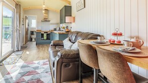 A cozy living area with a sofa, a dining table with a teapot, and a kitchen in the background. - Ponsford Ponds - Goosedown Lodge (Knowle, near Cullompton)