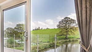 A wooden deck with a glass door leading to a scenic view of a river, grassy fields, and trees. - Ponsford Ponds - Goosedown Lodge (Knowle, near Cullompton)
