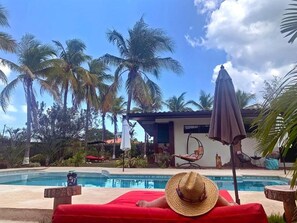 Un área junto a la piscina con una silla de relax roja, un sombrero de paja y una hamaca colgante.