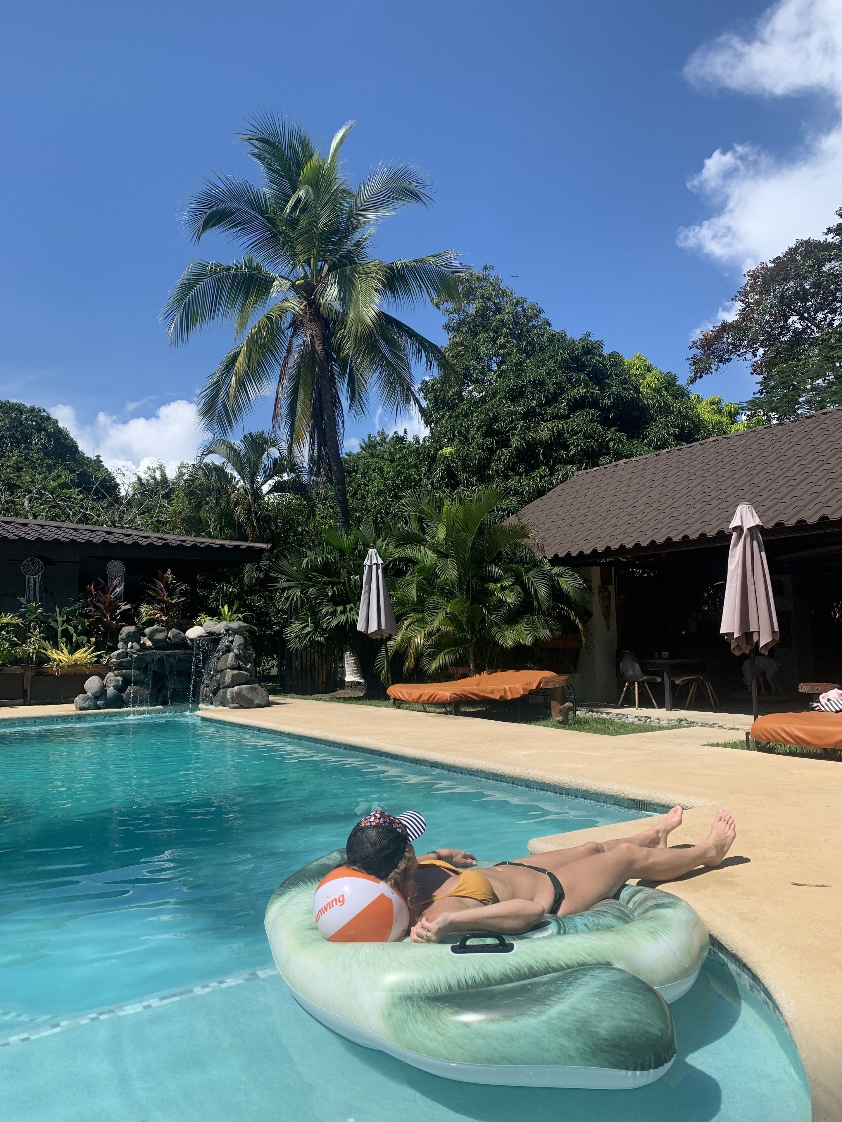 A pool area with a person lying on a float, surrounded by palm trees and a small waterfall.