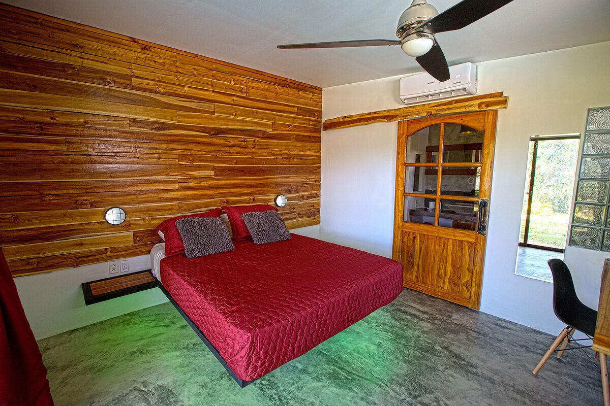 Quarto com cabeceira de madeira, colcha vermelha, ventoinha no teto e guarda-roupa de madeira.
