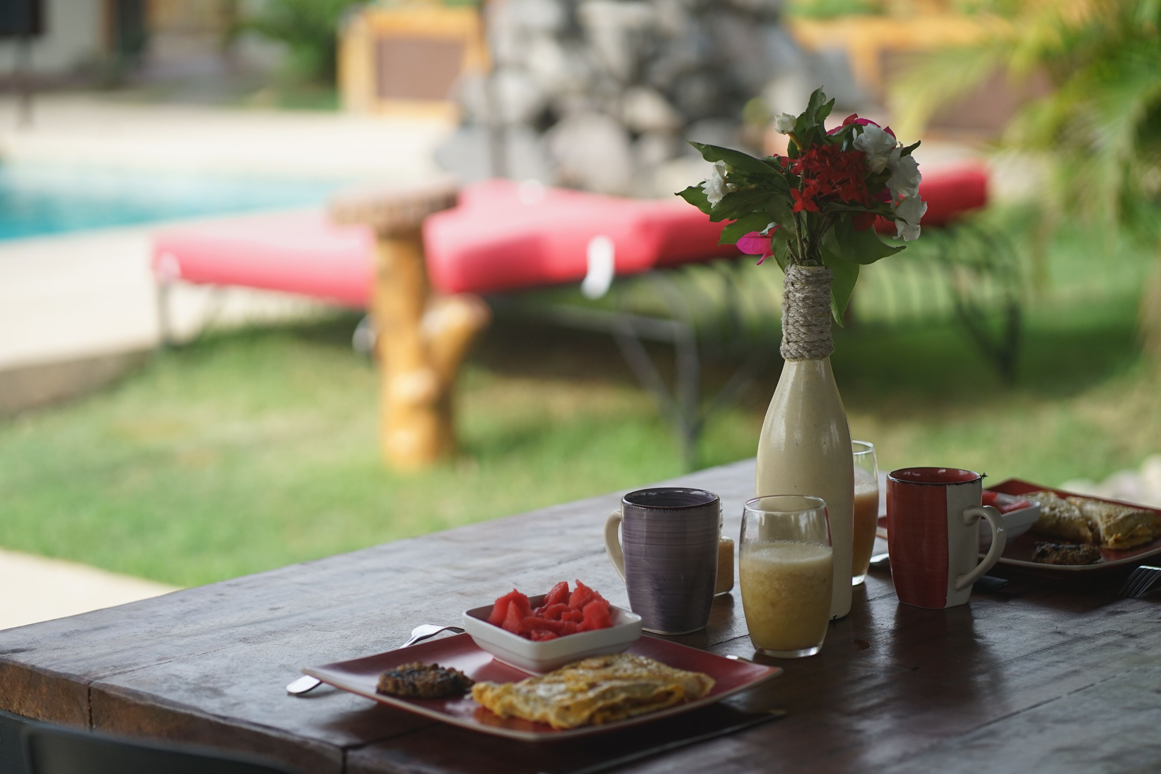 A dining table with breakfast items, a vase with flowers, and a pool in the background.