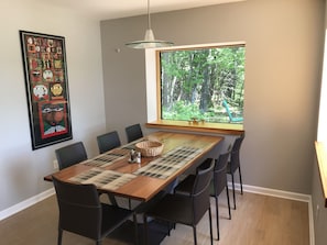 Un comedor con una mesa de madera, sillas negras, un protector de mesa tejido y un ventanal con vistas al bosque.