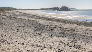 Een zanderig strand met een golvende kustlijn, rotsformaties en heuvels in de verte.
