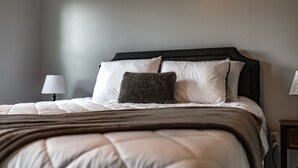 Ein ordentlich bezogenes Bett mit dunkelbrauner Kopfteil, weißen und dunkelgrauen Kissen sowie einer hellen Steude.