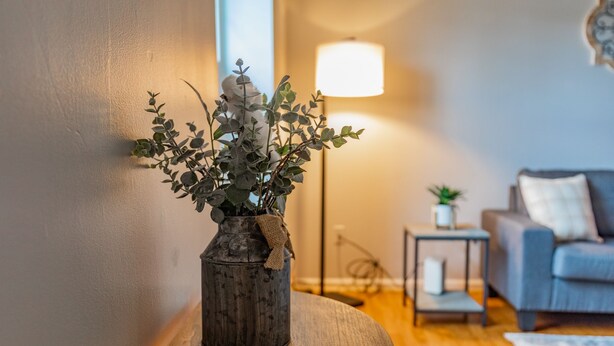 Ein Wohnzimmer mit einem runden Holztisch, einer Vase mit Grünpflanzen, einer Lampe und einem Sofa.