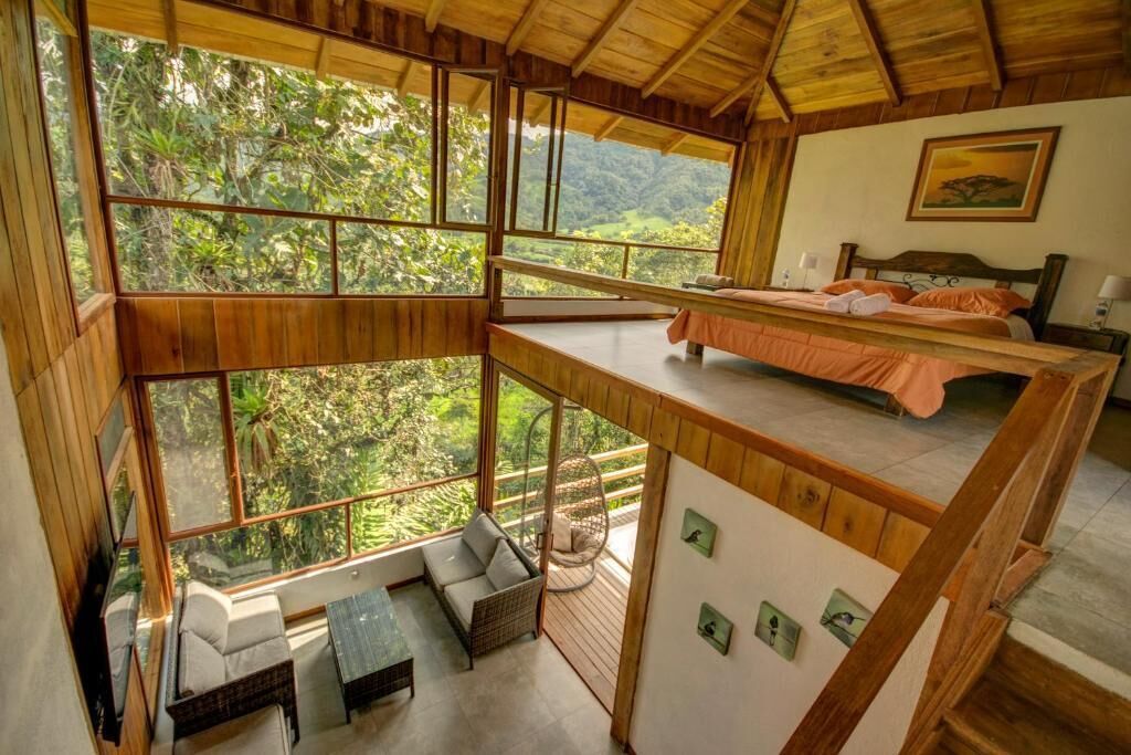 A high-ceilinged room with a bed, a sofa, and a dining table, all with large windows offering a view of the forest.