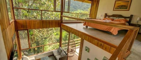 A high-ceilinged room with a bed, a sofa, and a dining table, all with large windows offering a view of the forest.
