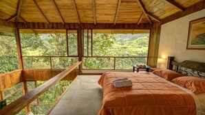Un dormitorio con un ventanal amplio que da a exuberante follaje, techo de madera y cama con colcha naranja.